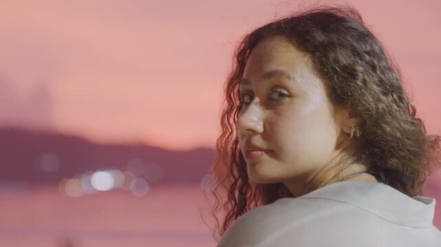 Woman portrait on island shore at sunset turning gaze toward camera soft profile curl hair gentle smile coastal twilight lights intimate candid portrait mood reflective storyteller on vacation