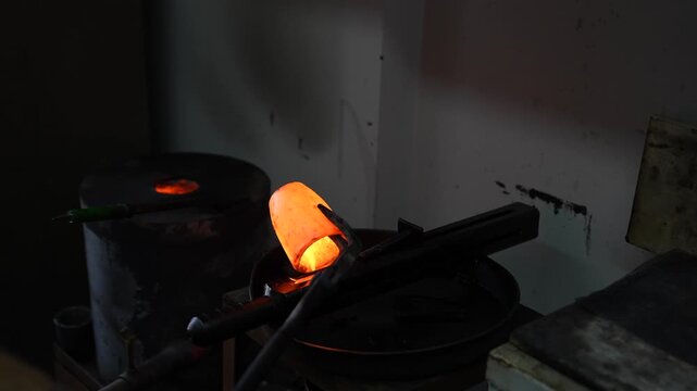 Goldsmith handling a crucible filled with molten gold using tongs after furnace melting. Shows goldsmithing process, high-temperature metalwork and casting preparation in workshop.