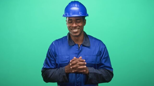 Man in blue hardhat and coveralls with clasped hands smiling on a green studio set, gesturing openly; pride duty.