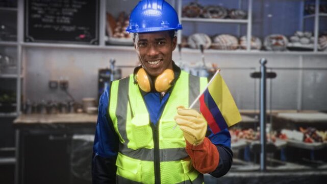 African american builder in blue hardhat and neon hi vis vest holds a small colombian flag with gloved hand and smiles, wearing yellow ear muffs in a building kitchen; pride solidarity.