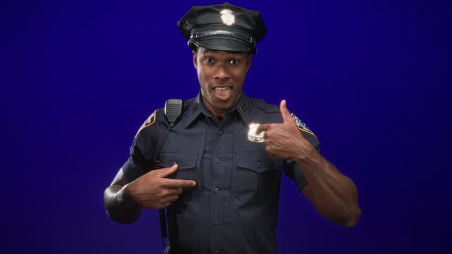 Man police officer points to badge with hand and thumbs at chest in studio portrait, wearing uniform and cap, smiling directly at camera; pride.