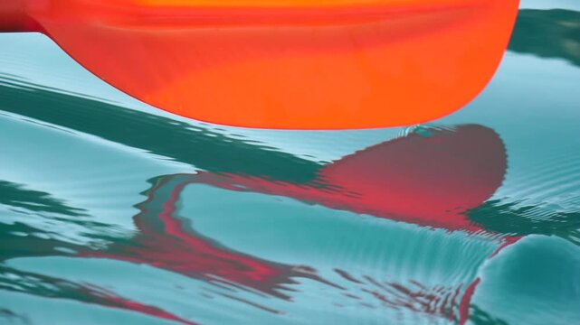 Paddle water surface, bright orange paddle skimming calm blue-green water, creating ripples and reflections