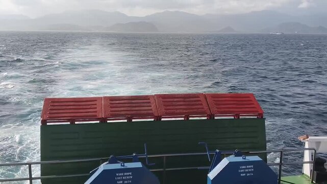 A dynamic high-angle shot from the aft (stern) of a large passenger and vehicle ferry, showcasing the powerful wake cutting through the deep blue ocean towards a majestic, distant misty mountain range