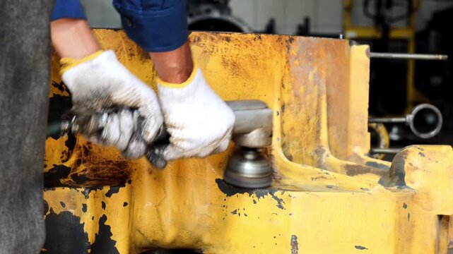 A technician in white safety gloves uses a pneumatic wire brush tool to strip old paint and rust from a heavy yellow industrial metal machinery component in a workshop.