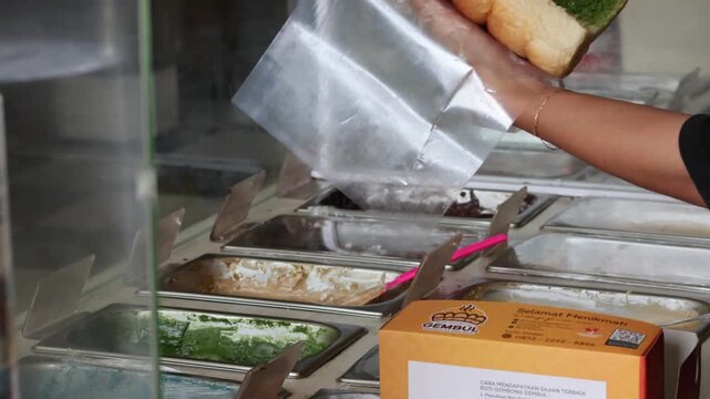 ​A street food vendor wearing plastic gloves uses a green spatula to spread jam inside a soft pull-bread roll in a shop, preparing a fresh snack for a customer with hygienic food handling.