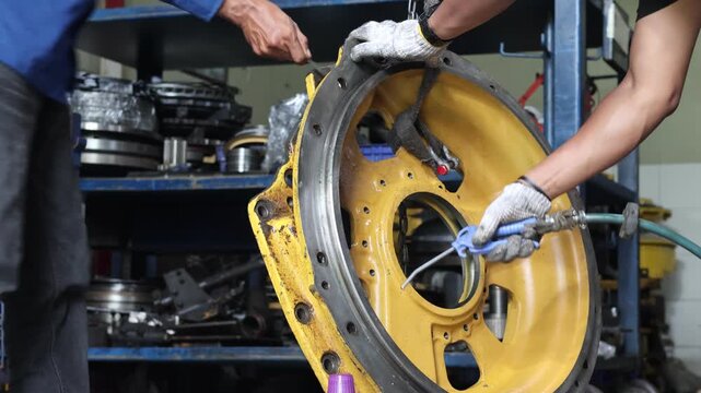 A mechanic uses a high-pressure air gun to blow dust and debris off a yellow industrial transmission cover in a workshop, ensuring a clean surface for reassembly during machinery maintenance.