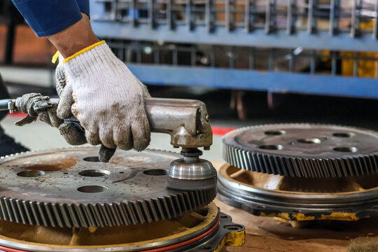 Close up of a mechanic using a pneumatic angle grinder to clean and polish a heavy industrial metal gear in a workshop for maintenance and repair.