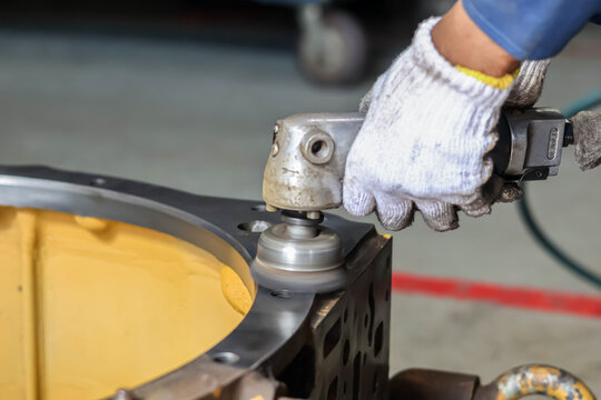 A technician uses a pneumatic orbital sander to polish the metal surface of a heavy yellow industrial component in a workshop, ensuring a smooth finish during a precision machinery overhaul.