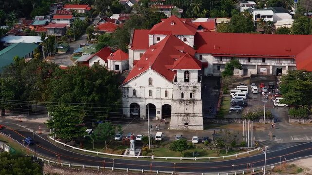 Front aerial view of Baclayon Church facade and bell tower beside curved road in historic town Bohol Philippines, drone