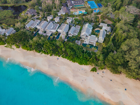 Aerial view of turquoise waters meeting the sandy shore, fringed by lush green trees and modern white buildings, Quatre Bornes, Mahe Island, Seychelles.
