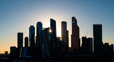Fototapeta premium Dark silhouetted skyscrapers against bright blue sky with sun flare and city skyline silhouettes