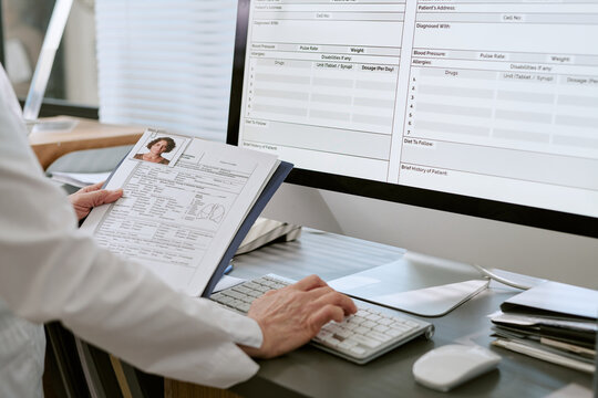 Middle aged healthcare worker reviewing patient file and entering medical data into electronic records on desktop monitor, supporting clinical documentation and hospital administration use