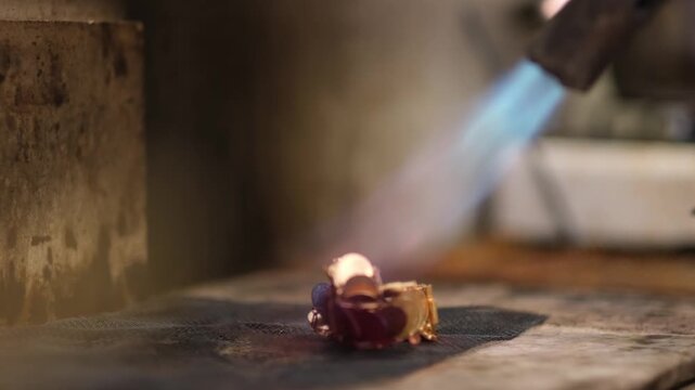 Close-up of a gold jewelry piece being soldered with a torch flame. Highlights traditional craftsmanship, heat processing and precision metalworking in a professional jewelry workshop.