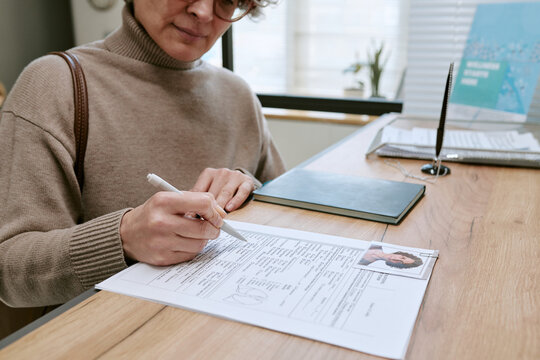 Middle aged Caucasian woman signing application form at office desk, completing identity verification and paperwork. Suitable for HR recruitment, onboarding, visa, banking content