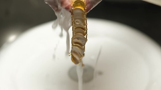 Goldsmith rinsing gold jewelry under running water after cleaning process. Shows goldsmithing workflow, surface purification and final preparation stage in traditional jewelry workshop.