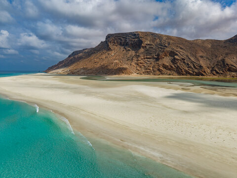 Aerial view of a sun-kissed beach meeting the turquoise lagoon waters under a sky streaked with clouds, Detwah lagoon, Socotra, Yemen.