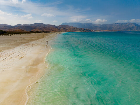 Aerial view of turquoise waters gently kiss the golden shores, a solitary figure graces the serene landscape against the backdrop of rugged mountains, Shoab beach, Socotra, Yemen.