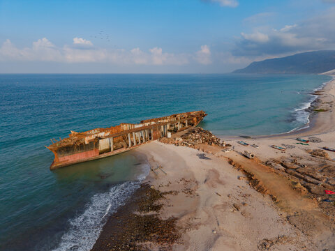 Aerial view of a rusted shipwreck, beached on the sandy shore, where the turquoise sea meets the rugged coastline, Ararhin, Yemen.
