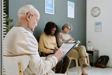 Naklejka premium Senior Caucasian man filling medical intake form in clinic waiting room while diverse patients using clipboard and smartphone. Useful for healthcare access, patient registration, insurance marketing