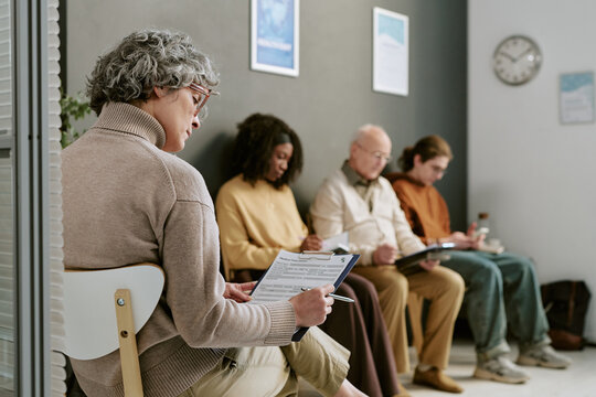 Senior Caucasian woman reviewing intake form in clinic waiting room while diverse patients completing paperwork, representing healthcare access, patient registration, insurance marketing