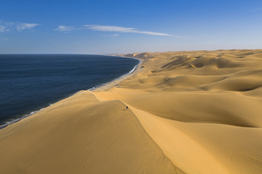 Aerial view of golden dunes meeting the deep blue ocean, creating a striking contrast of warm and cool tones, Walvis Bay, Namibia.