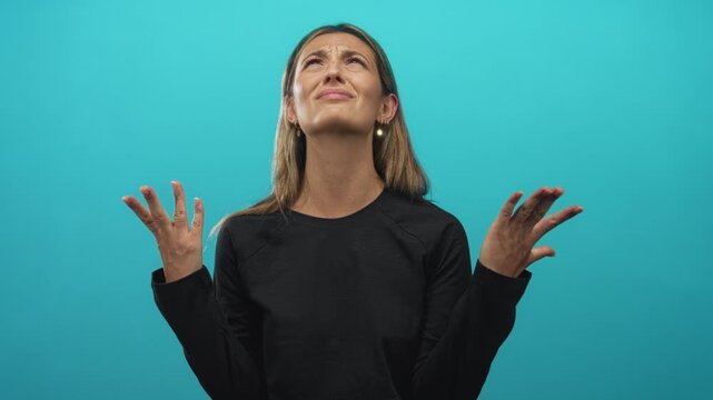 Young blonde woman raising hands in a shrug gesture, squinting and looking up in turquoise studio; frustration moment.