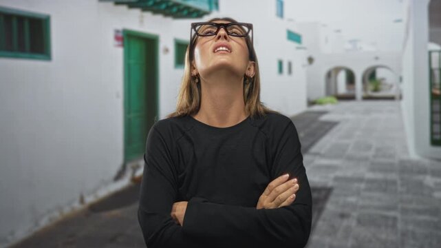 Young woman covering ears with hands and oversized glasses on a sunlit whitewashed street; frustration.