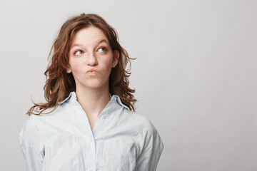 Fototapeta premium Young woman with curly hair in white shirt looks sideways with thoughtful expression on light background. Concept for doubt, decision making, and contemplation.