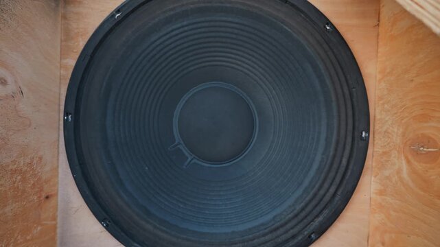 A large black subwoofer speaker cone sits inside a wooden acoustic cabinet. The corrugated paper diffuser vibrates heavily to produce deep bass frequencies and powerful audio.