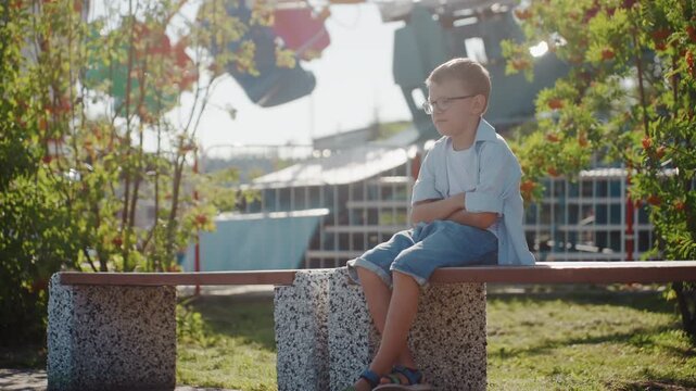Young white boy sitting bench park weekend family outing near amusement rides, patterned bag beside him, late afternoon sunlight, pensive expression, casual denim shorts, relaxed quiet moment