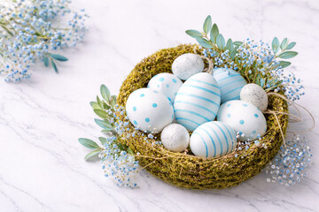 Obraz premium Elegant Easter flat lay with blue striped and polka dot eggs in moss nest, delicate blue flowers and fresh leaves on marble background, soft pastel spring decor, stylish seasonal stock image.