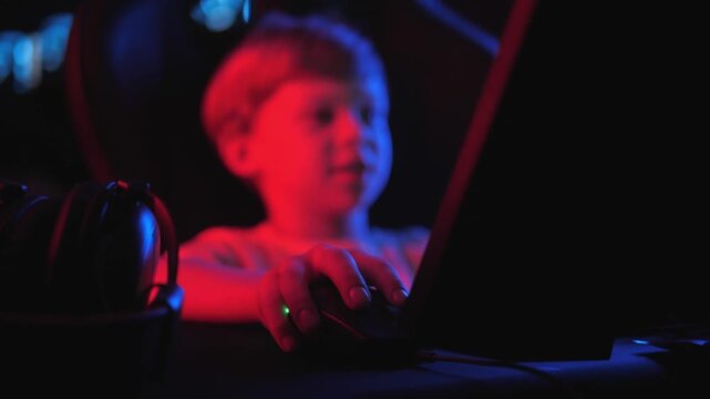 Close-up of a little boy playing a video game on a computer at night. Focus on the hand on the computer mouse. The player presses the computer mouse button.