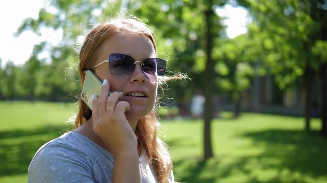 A picture of a contented lady wearing sunglasses, conversing on a sunny day in a verdant park. The image is unwavering. The camera remains steady.