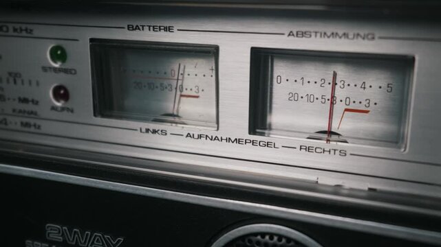 Illuminated analog VU meters indicate recording levels on a vintage stereo deck. Red needles fluctuate across dual metallic dials featuring retro German text. Classic audio equipment details.
