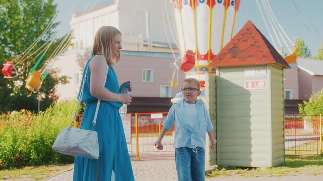 White mother and son walking past colorful swing ride in sunny park weekend, mother wearing blue dress and carrying white bag, boy smiling and pointing, gentle pace, carnival atmosphere, green shrubs