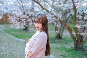 Fototapeta premium Young woman with long brown hair and bangs standing in a grassy area with blooming cherry blossom trees in the background.