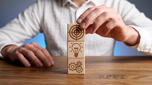 A man in a white shirt sits at a wooden table stacking wooden blocks with icons of gears, a light bulb, and a target to represent business strategy, planning, and goal achievement.