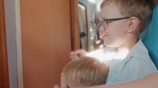 White brothers laugh at mirror, older sibling in glasses comforting younger child with hand on shoulder as sunlight glints on curved glass, candid indoor family moment full of amusement and warmth