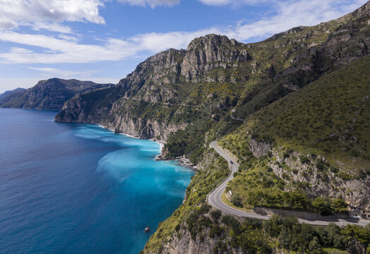 Aerial view of turquoise waters meet rugged cliffs along a winding coastal road, a symphony of blues and greens under a vast sky, Amalfi, Campania, Italy.