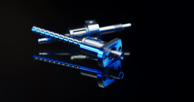 Two industrial metallic ball screw spindles with blue light illumination on a black reflective surface