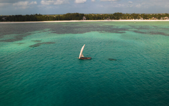 Aerial view of a traditional dhow sailing on turquoise waters, contrasting with the white sandy shore and the lush green trees along the coast, Paje, Unguja South Region, Tanzania.
