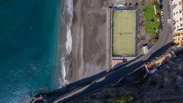 Aerial view of the dark sandy beach meeting the turquoise ocean next to a tennis court and colorful buildings, Minori, Salerno Italy