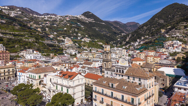 Aerial view of sun-kissed buildings with terracotta roofs cascade down the slopes towards the town square, nestled against the majestic mountains, Minori, Campania, Italy.
