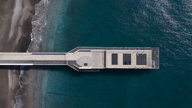 Aerial view of a pier extending into the tranquil, deep blue sea where the dark sand meets the foamy waves, Minori, Salerno Italy