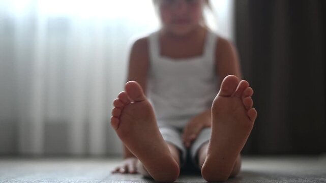 Close-up of little girl&rsquo;s bare feet while sitting on the floor. The soft focus emphasizes innocence and calmness, capturing a quiet childhood moment in a cozy home environment.