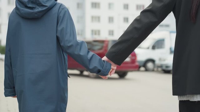 Child holding adult hand preparing school on urban street near parked cars blue coat hood up adult in dark coat escorting child across sidewalk gentle grip quiet morning focus on hands emotional bond