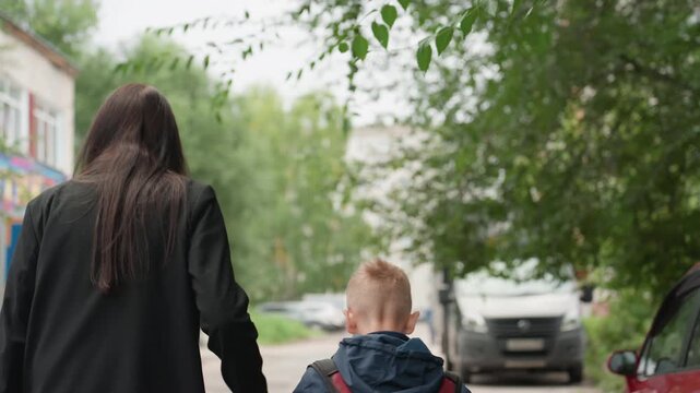 White mother holding son hand school, colorful facade and mural, treelined path, backpacks and cars, steady pace, supportive grip, warm morning light, emotional reassurance, preparing for class