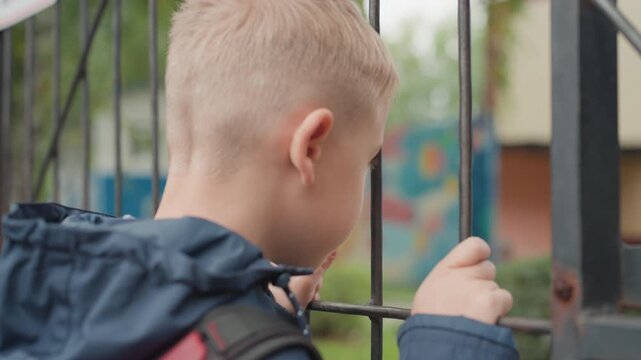 Young boy at school gate gripping fence, backpack and navy jacket, peering into schoolyard with hesitant posture, gripping rusty metal bars, quiet suburban campus, soft overcast light, shallow focus