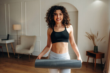 Obraz premium Young woman with curly hair in black sports bra holds yoga mat in modern living room with wooden floor and minimalistic furniture in the background