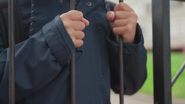 Young boy at school gate preparing bag. Closeup of hands gripping metal bars, navy jacket and hood, suburban school building in background, morning routine, hesitant expression, quiet anticipation